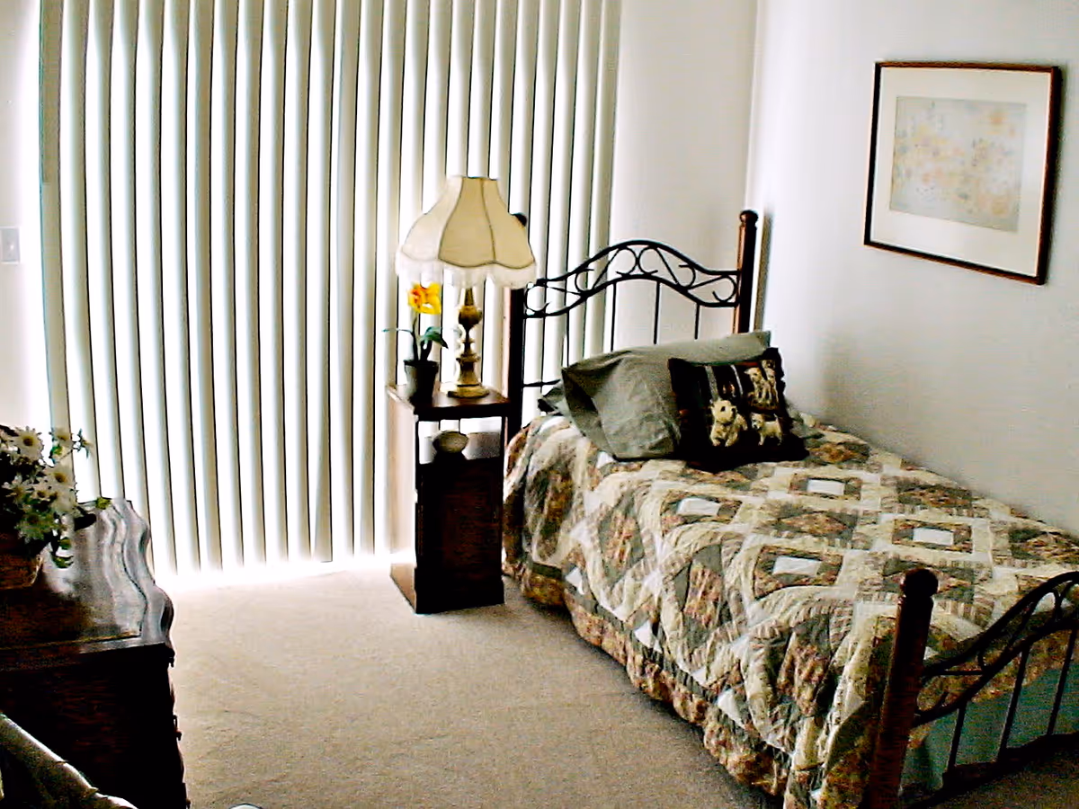 A small bedroom with a single bed covered in a patterned quilt. There are two pillows on the bed, one with a dog design. Next to the bed is a wooden nightstand with a vintage-style lamp and a small potted plant. Vertical blinds cover a large window or sliding door, allowing light to filter into the room. A framed picture hangs on the wall above the bed, and a wooden dresser with a vase of flowers is partially visible on the left side.