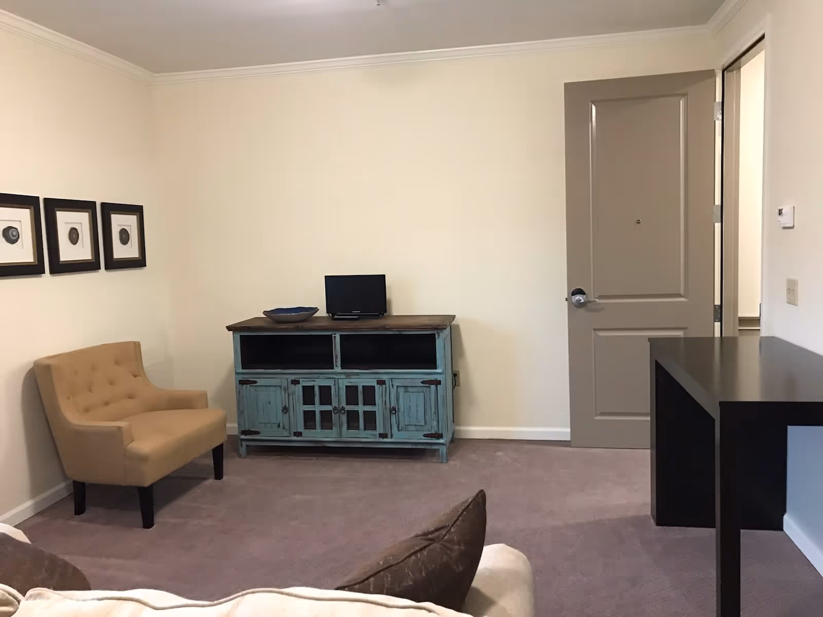 Small living room with a beige armchair, teal TV console with a small TV, a desk, and a closed door against a neutral wall.