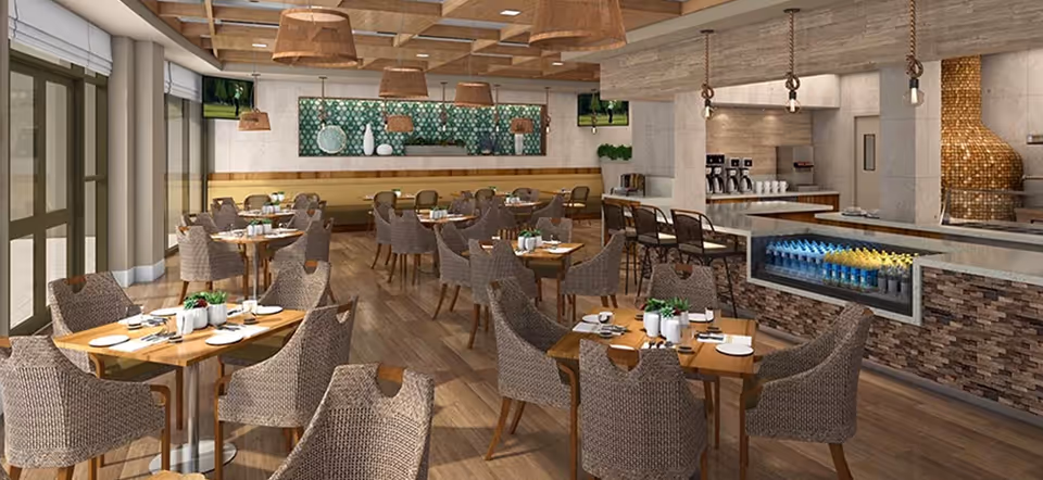 A spacious dining area with multiple wooden tables and wicker chairs arranged neatly. The room features large windows on one side allowing natural light to enter. There is a counter area with bar stools and a display of bottled drinks underneath. The ceiling has wooden beams with hanging light fixtures, and the walls are decorated with green patterned artwork and shelves with decorative items.