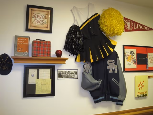 A wall decorated with nostalgic school memorabilia including a framed 'School Days' sign, a Brownie Scout handbook, a red plaid lunchbox, a red apple, a black and yellow cheerleading outfit with matching pom-poms, a letterman jacket with the letters 'SM', a Lincoln pennant, and various framed documents and photos.
