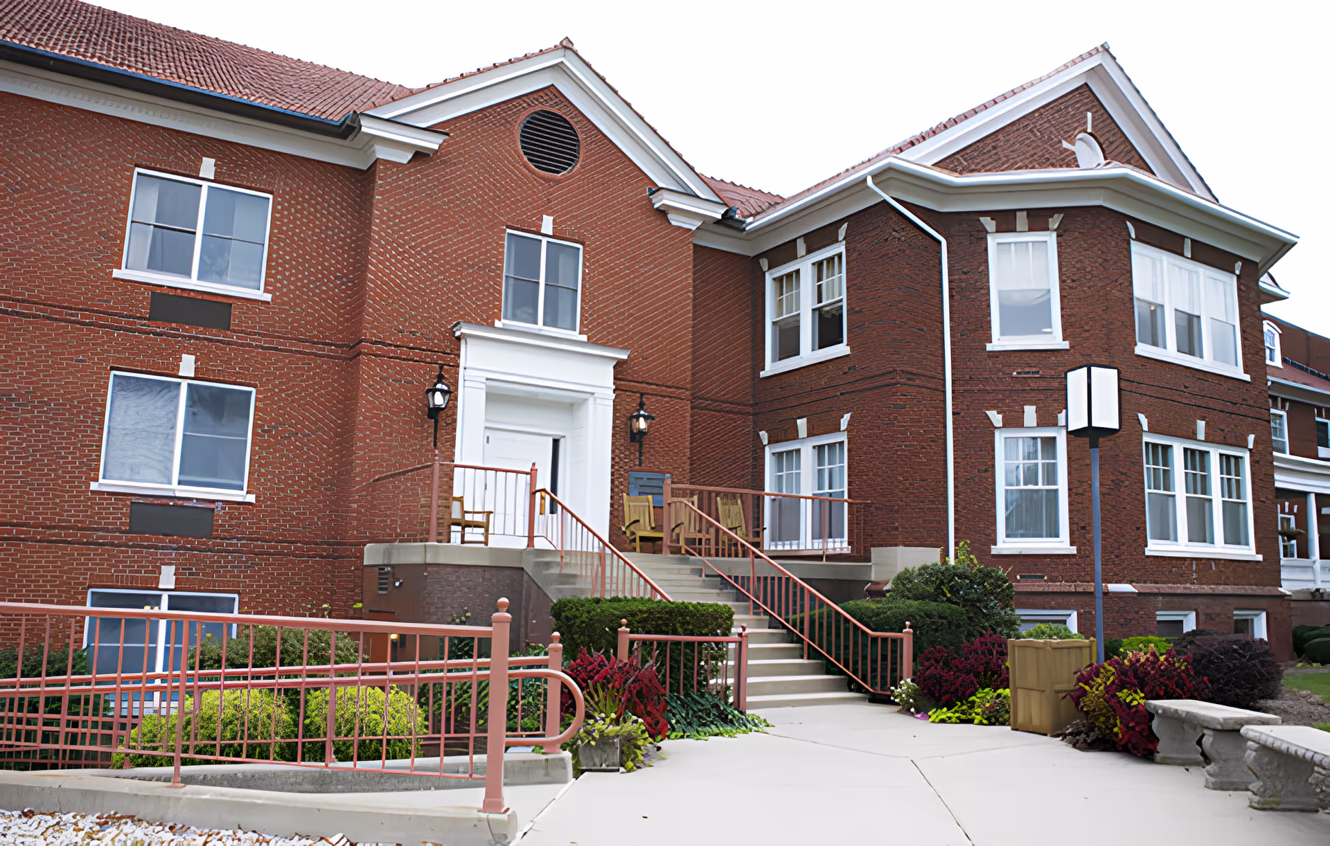 Exterior view of a red brick building with white window frames and a tiled roof. There is a staircase leading up to a white door entrance with two chairs on the porch. The area is landscaped with bushes and flowers, and there is a paved walkway and a ramp with railings.