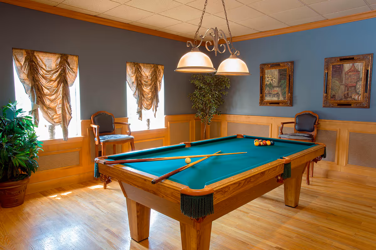A well-lit game room with a wooden pool table covered in green felt. Two pool cues and a white cue ball are on the table, with a rack of billiard balls at one end. The room has wooden flooring, two windows with ornate curtains, two wooden chairs with dark cushions, a potted plant, and two framed paintings on the blue-gray walls. A decorative hanging light fixture with two lamps illuminates the table.