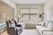 A bright and cozy living room area with a large window letting in natural light. The room features a comfortable dark recliner chair on the left, a white sofa on the right, a white side table with a lamp, and a bed partially visible through an open doorway on the left side.