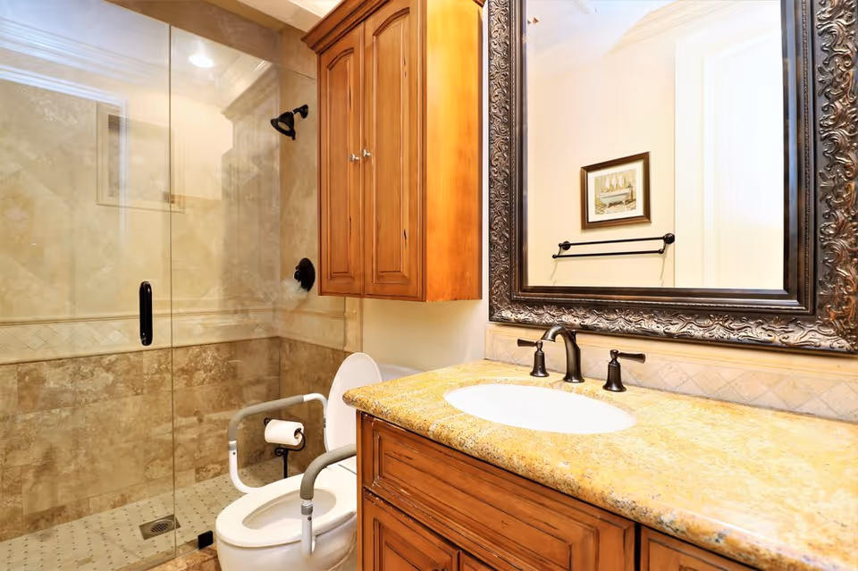 Well-appointed bathroom with a glass-enclosed shower, wooden vanity with granite countertop and a toilet fitted with grab bars.