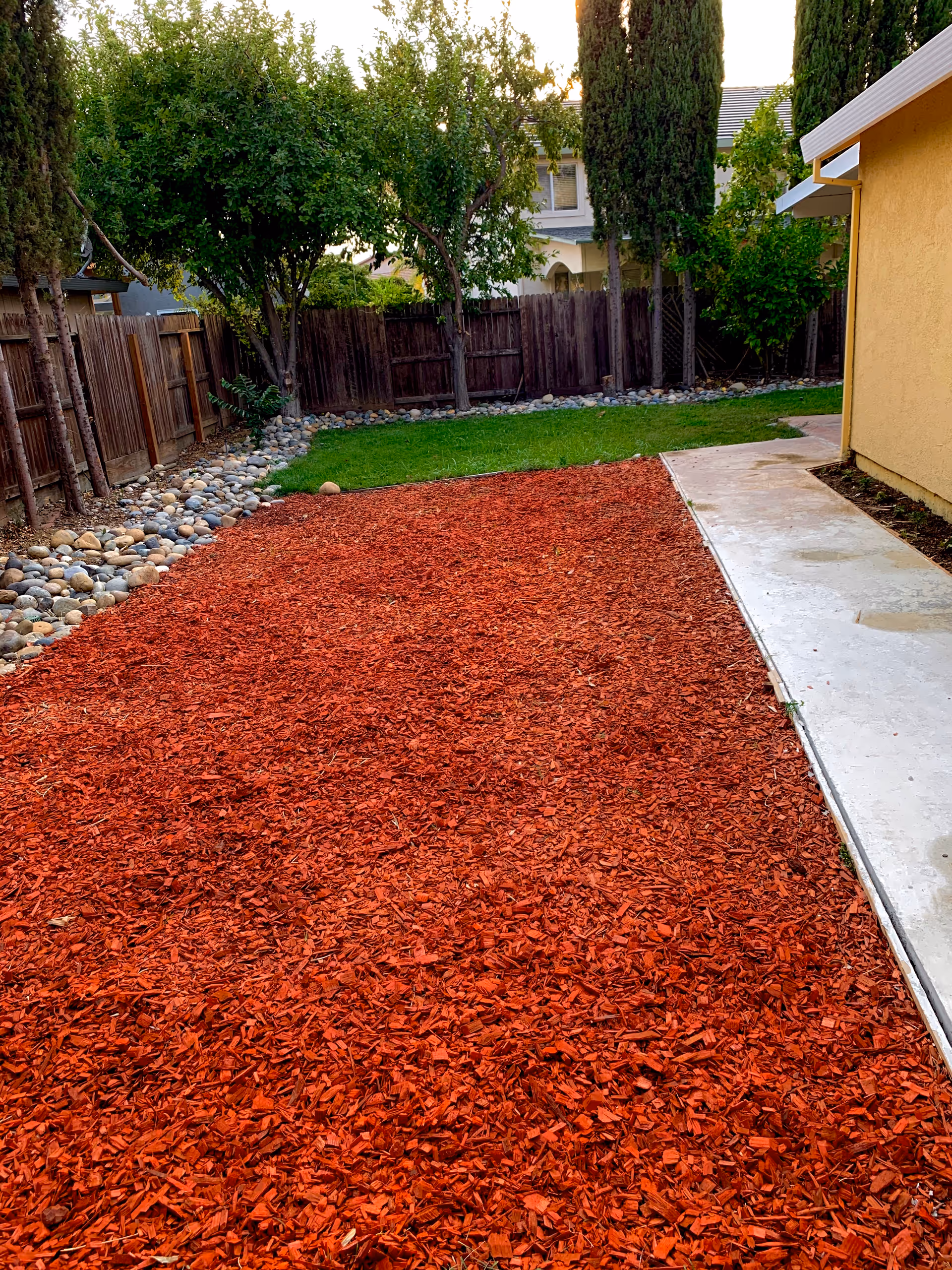 Outdoor area with a red wood chip ground cover, a concrete walkway on the right, green grass and trees along a wooden fence in the background, adjacent to a yellow building.
