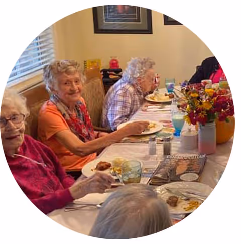 A group of elderly women sitting around a dining table enjoying a meal together in a well-lit room with a window and framed pictures on the wall. The table is set with plates of food, glasses, and a vase of colorful flowers.