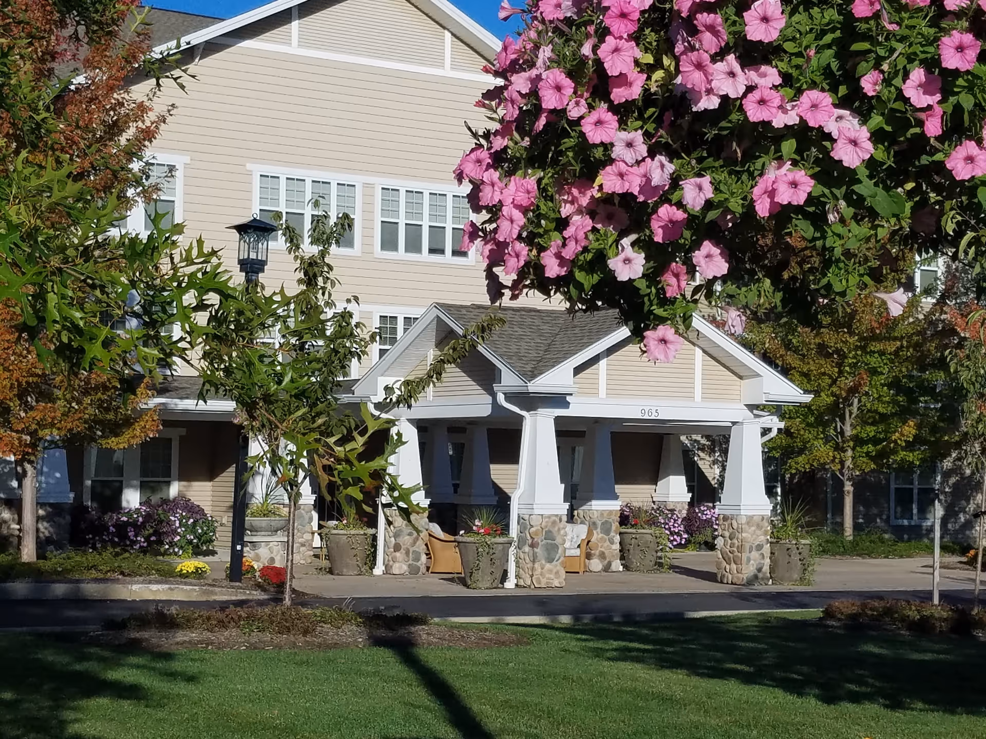 Front exterior view of a beige senior living facility building with white trim, a covered entrance supported by stone pillars, surrounded by green trees and pink flowers in bloom.