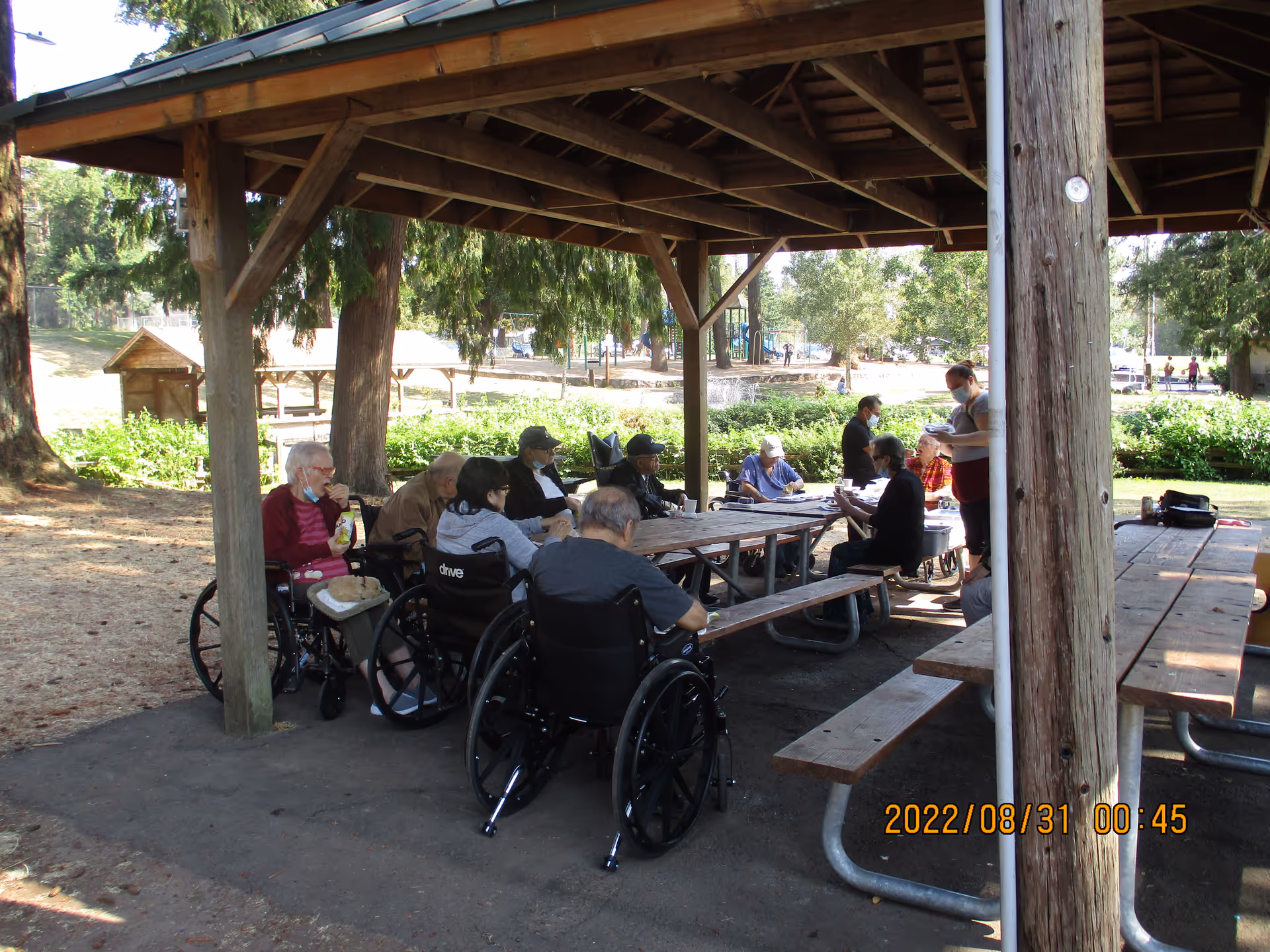 A group of elderly people, some in wheelchairs, sitting around picnic tables under a wooden pavilion in an outdoor park-like setting. A caregiver wearing a mask is standing and interacting with the group. Trees and greenery surround the area.