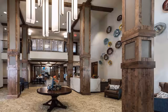 Spacious senior living facility interior with tall wooden columns, a round wooden table with decorative items, comfortable armchairs, and a wall adorned with colorful decorative plates. The area includes a dining space in the background and a second-floor balcony overlooking the main floor.