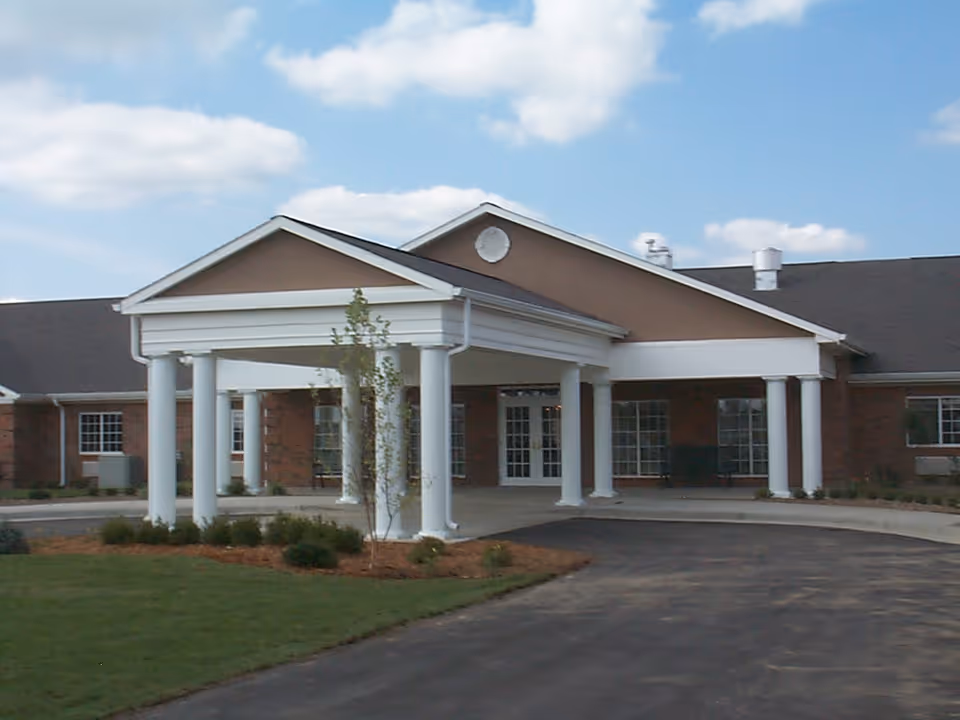 Front exterior view of Waterford Place Health Campus building with a covered entrance supported by white columns, a driveway, and some landscaping including grass and small bushes under a partly cloudy sky.
