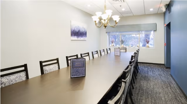 Long dark dining table with many chairs in a bright room featuring a chandelier, window, and wall art.