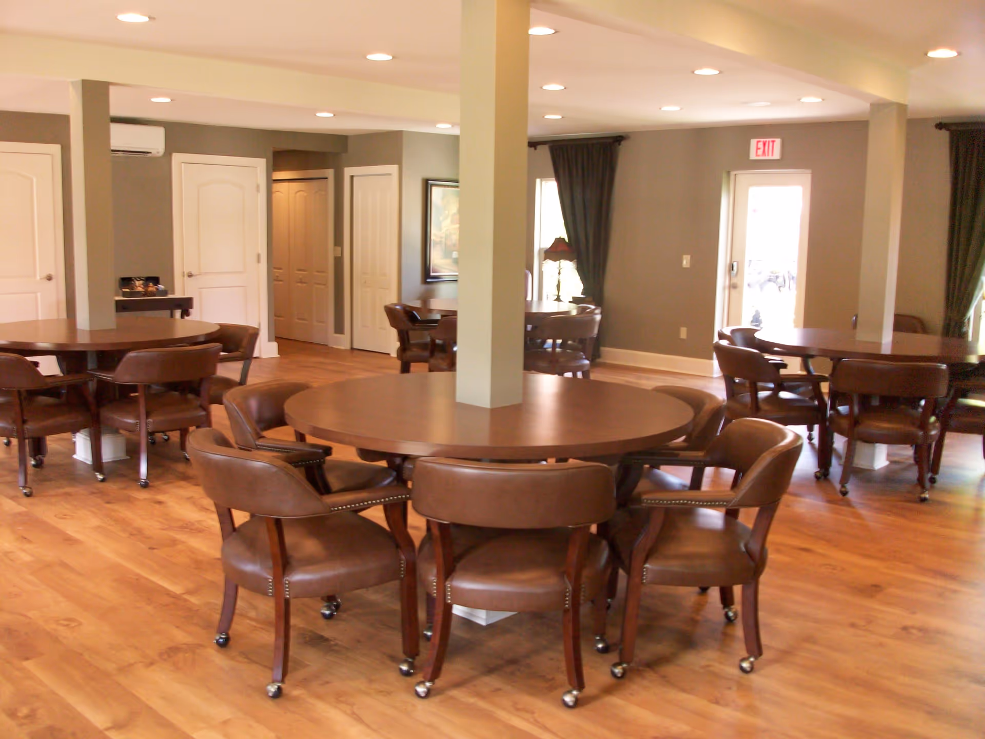 Interior view of a senior day program common area with multiple round wooden tables surrounded by brown cushioned chairs on wheels, hardwood flooring, beige walls, and recessed ceiling lights. There are several doors and windows with dark curtains, and an exit sign above a door in the background.