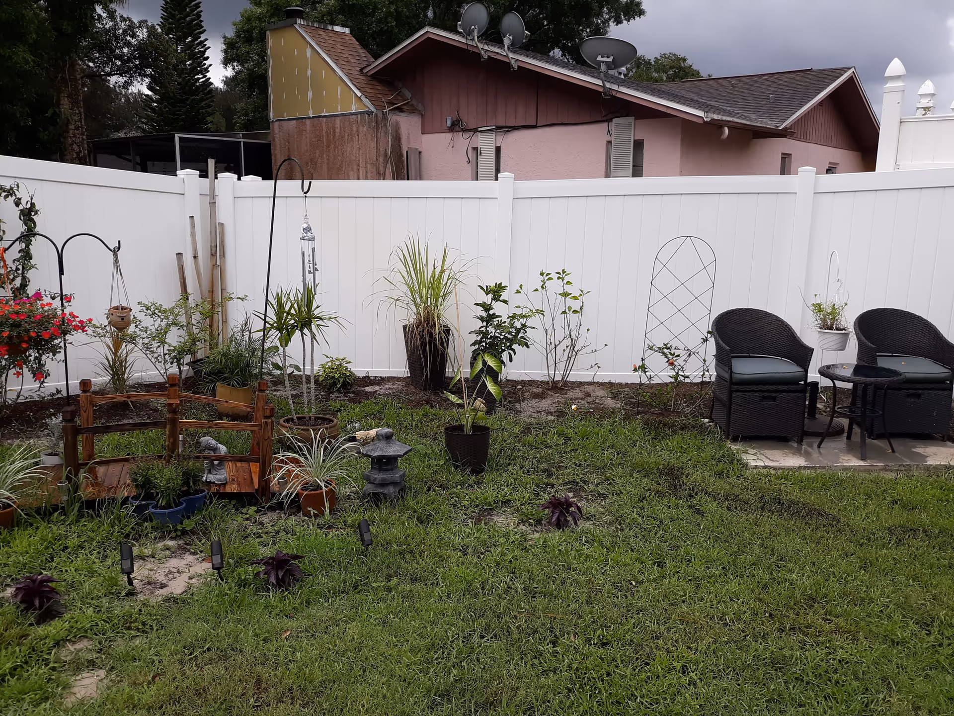 A small garden area with various potted plants and garden decorations in front of a white fence. There are two black wicker chairs with cushions and a small glass-top table on a concrete patio section to the right. In the background, there is a pink house with a brown roof and multiple satellite dishes on top.
