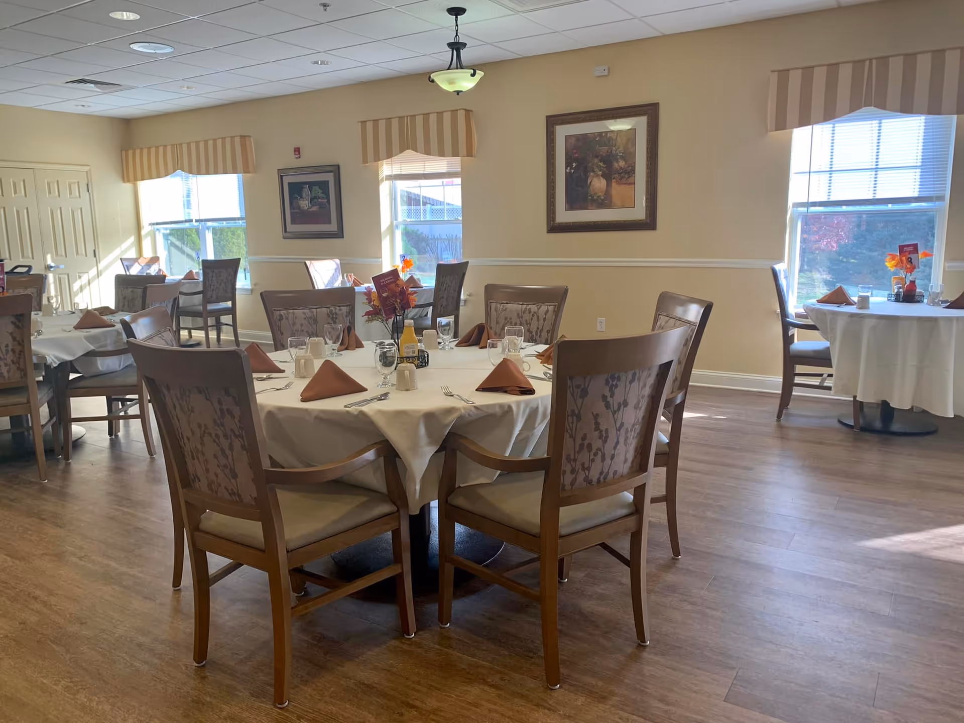 A well-lit dining room with round tables covered in white tablecloths, each set with brown napkins, glasses, and silverware. The room has wooden chairs with patterned upholstery, large windows with striped valances letting in natural light, and framed artwork on the walls. The floor is wooden, and there are small floral centerpieces on the tables.
