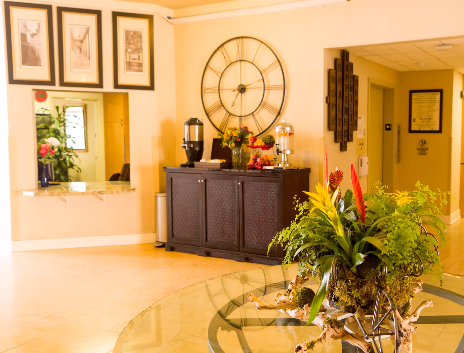 Interior view of a retirement village common area featuring a glass table with a decorative plant centerpiece in the foreground. In the background, there is a dark wooden cabinet with a large wall clock above it, a beverage dispenser, a fruit stand, and a vase with flowers. The walls are decorated with framed pictures and a wooden wall art piece. The floor is tiled and the lighting is warm.