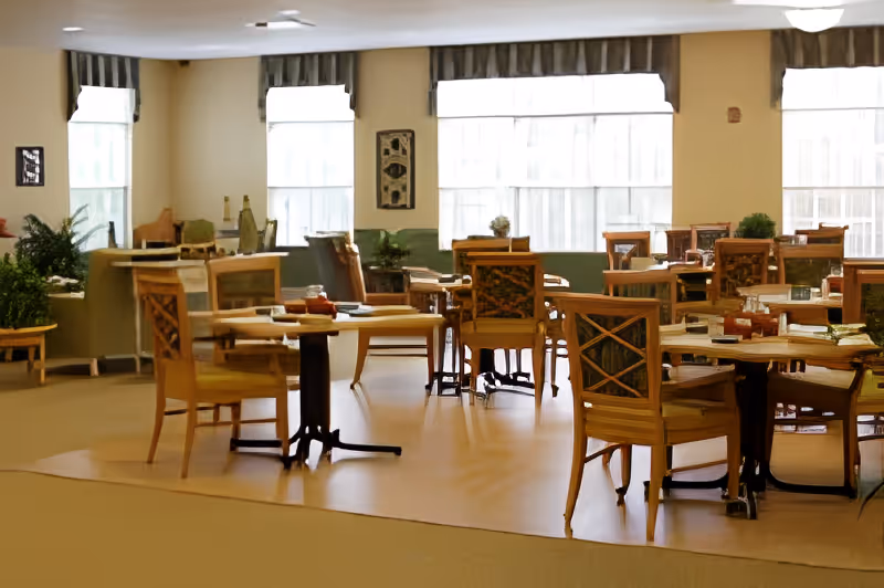 Bright communal dining room with multiple wooden tables and chairs arranged near large windows.