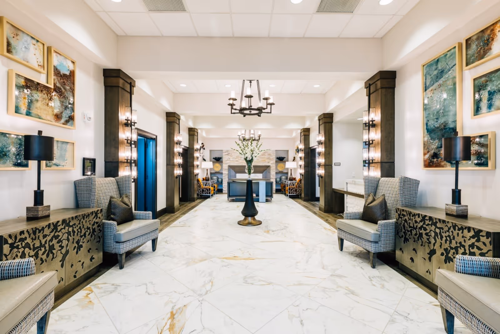 A spacious and elegant interior hallway with marble flooring, two patterned armchairs on each side, decorative cabinets with black lamps, abstract artwork on the walls, and a central round table with a vase of white flowers. The hallway is well-lit with wall sconces and chandeliers, featuring wooden columns and a cozy seating area with a fireplace at the far end.