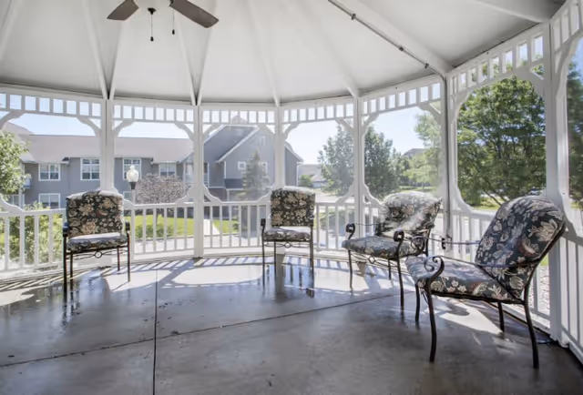 A bright, white gazebo with a ceiling fan and four cushioned metal chairs arranged inside. The gazebo overlooks a landscaped area with trees and residential buildings in the background under a clear sky.