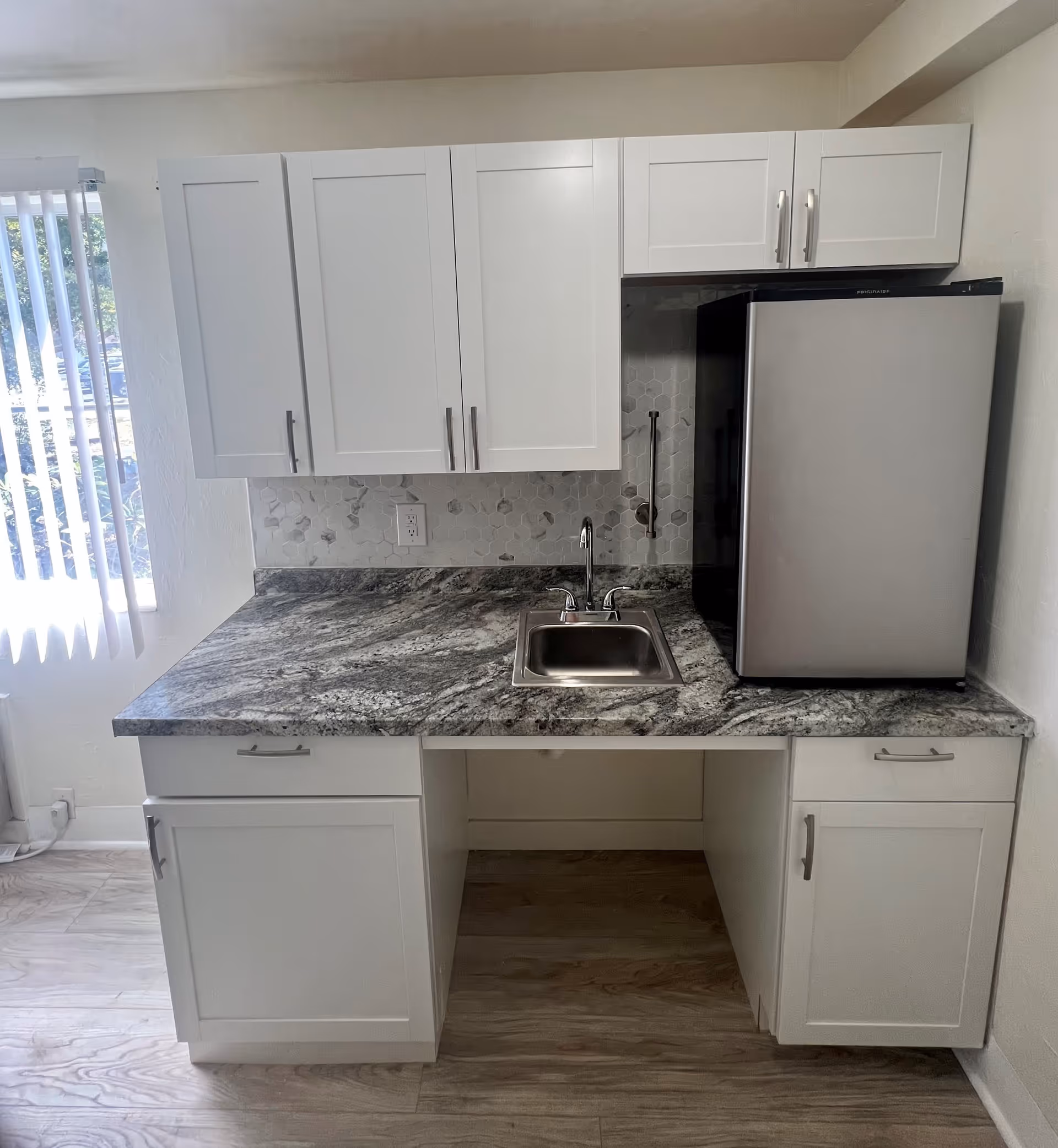 A small kitchen area with white cabinets, a gray and white marbled countertop, a stainless steel sink with a faucet, and a compact stainless steel refrigerator. There is a window with vertical blinds to the left, and the backsplash features hexagonal tiles in shades of white and gray.