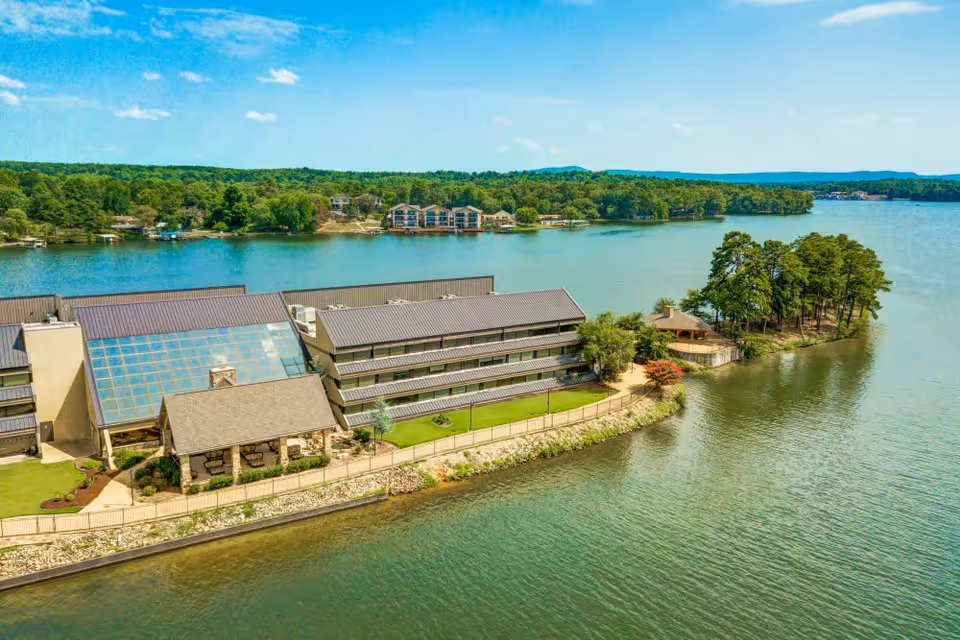 Aerial view of The Atrium at Serenity Pointe facility located on a waterfront with a large building featuring multiple floors and a glass atrium. The building is surrounded by water on three sides with a small tree-covered peninsula extending into the water. The background shows a forested area and a clear blue sky.