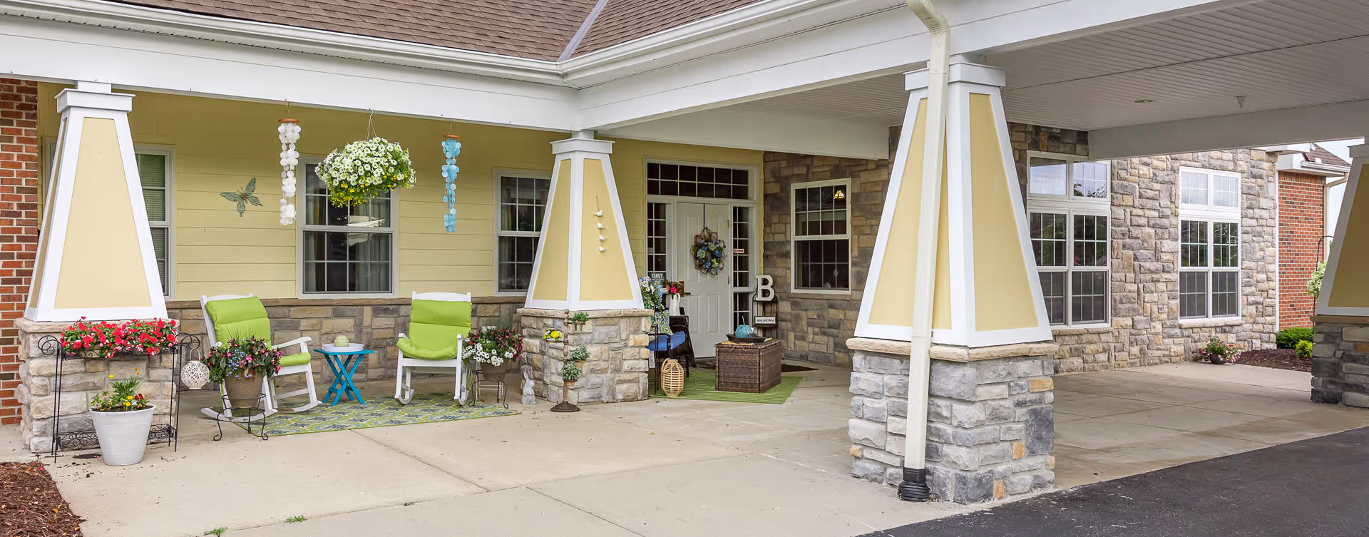 Covered outdoor patio area at a senior living facility with stone pillars, yellow siding, and a white door decorated with a wreath. There are two green cushioned chairs, a small blue table, potted plants, and hanging flower baskets. The area is clean and inviting with a concrete floor and a driveway visible on the right.