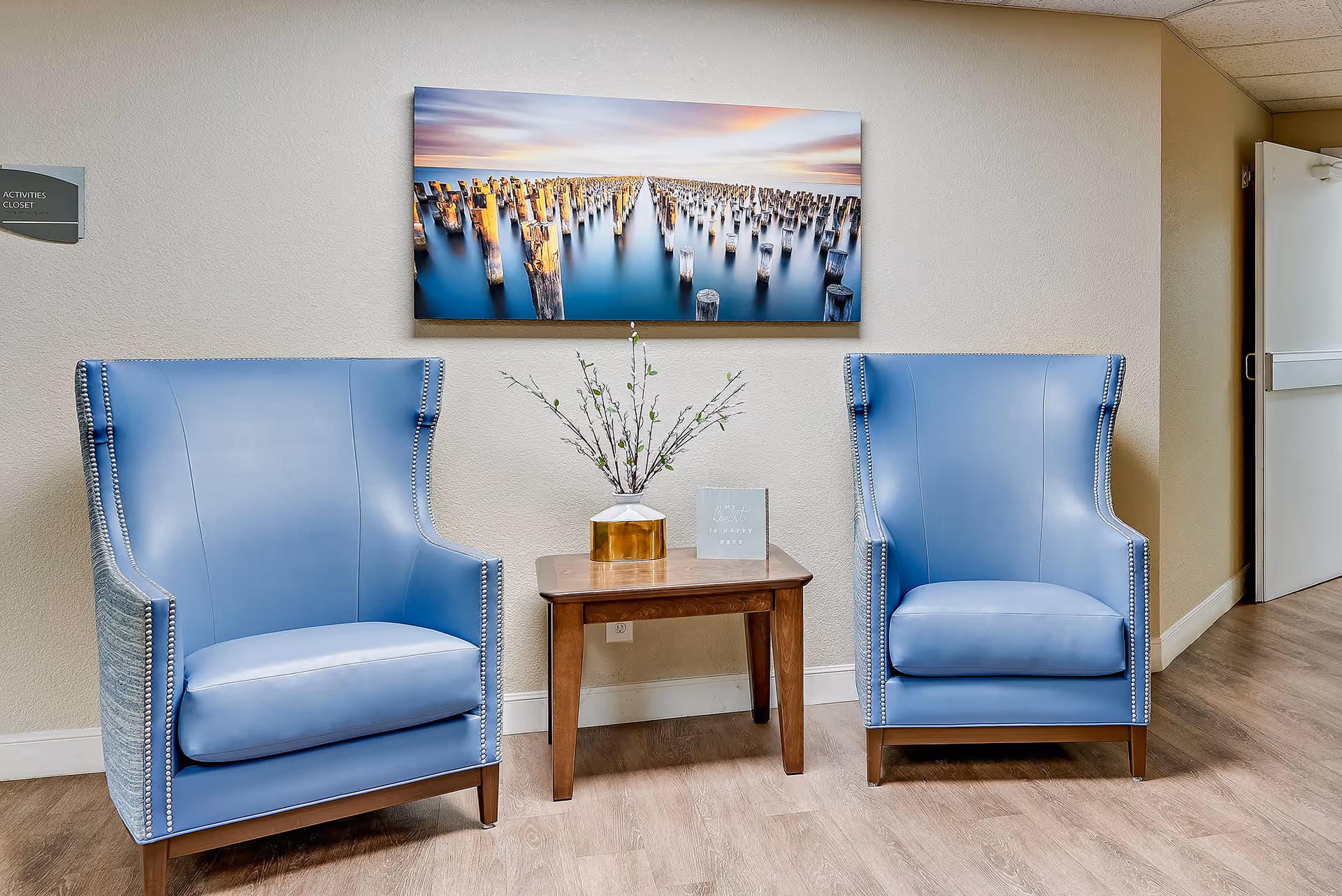 Two blue armchairs flanking a small wooden table with a vase and wall art above in a hallway seating area.