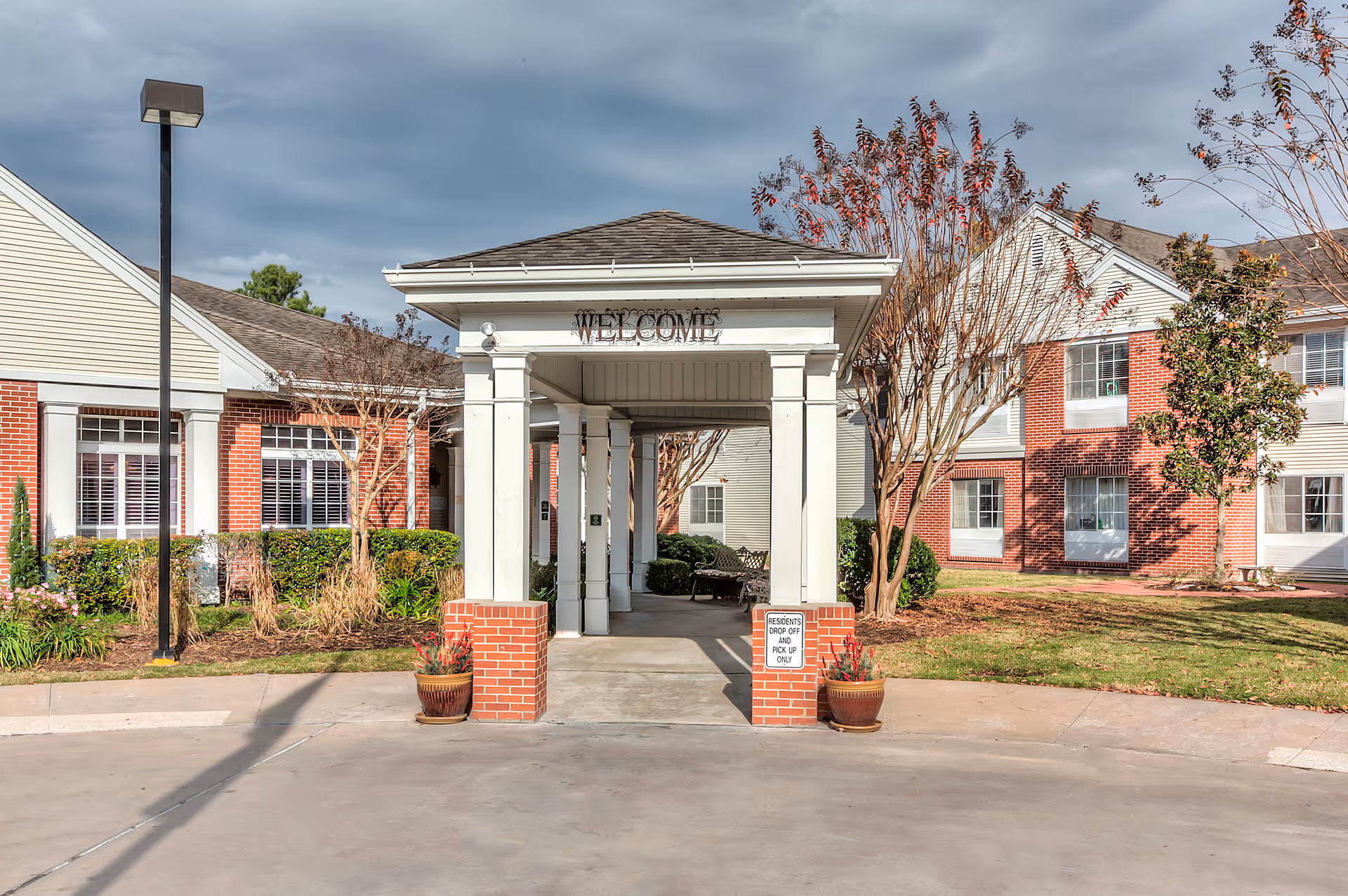 Entrance to a senior living facility with a covered drop-off area supported by white columns and brick bases. The building has red brick and beige siding with multiple windows. There are landscaped bushes and trees around the entrance, and a sign that reads 'Residents Drop Off Pick Up Only'. The sky is cloudy.