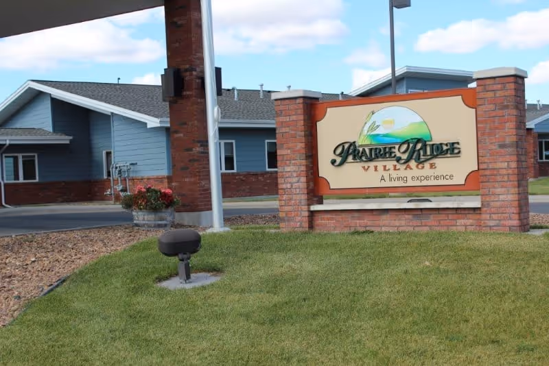 Outdoor view of Prairie Ridge Village facility entrance sign made of brick with a landscaped lawn and buildings in the background under a partly cloudy sky.