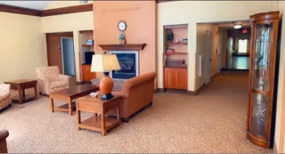 A cozy senior living facility common area with beige carpet and walls. The room features a brown leather sofa, two patterned armchairs, wooden side tables with lamps, a coffee table, and a fireplace with a clock and decorative items on the mantel. There is a hallway leading to other rooms and a glass display cabinet on the right side.