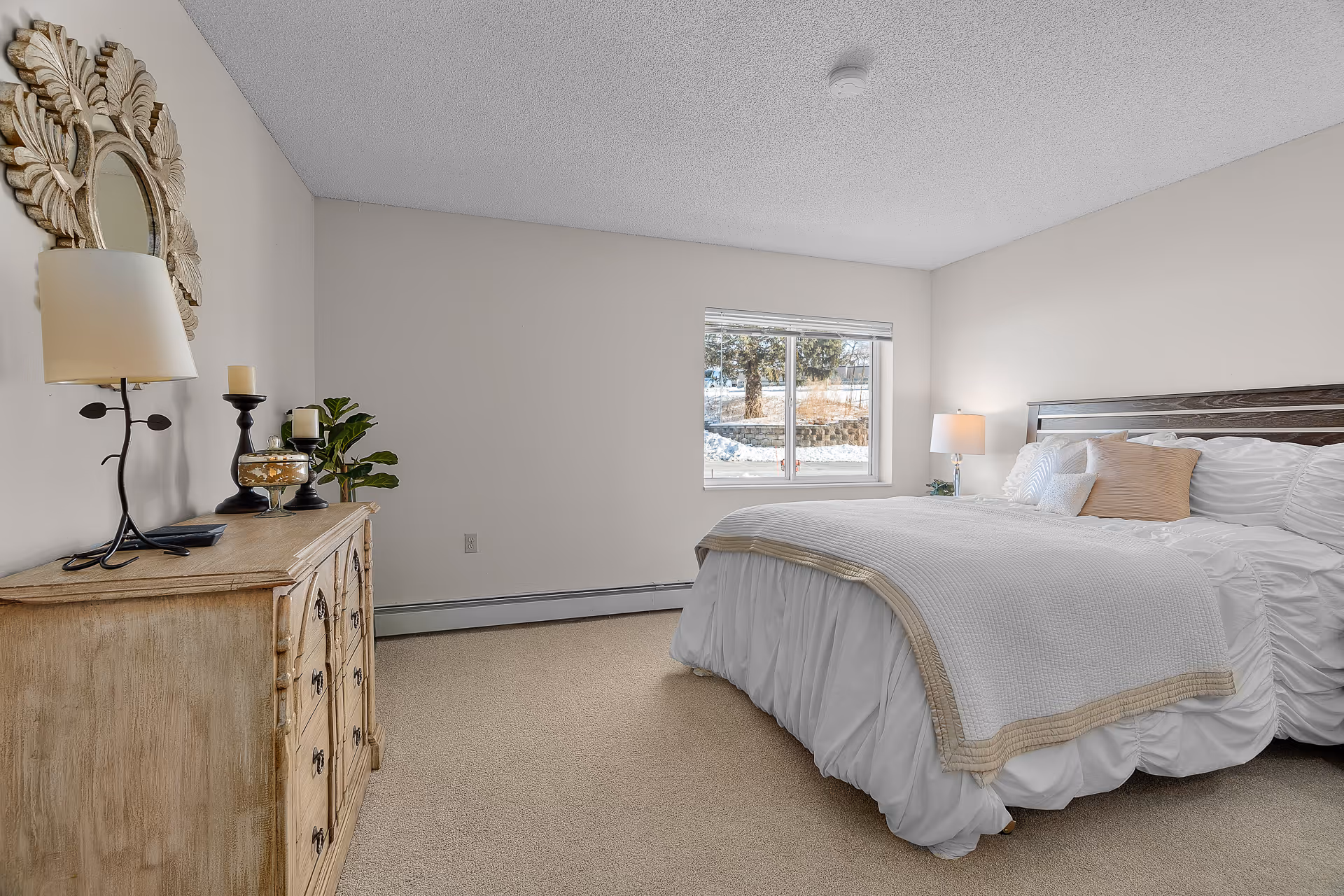 Bright, neutral bedroom with a made bed, wooden headboard, dresser topped with lamps and decor, and a window showing an outdoor view.