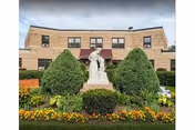 A beige brick building with multiple windows behind a garden featuring a white statue of a person holding a child, surrounded by green bushes and colorful flowers under a partly cloudy sky.