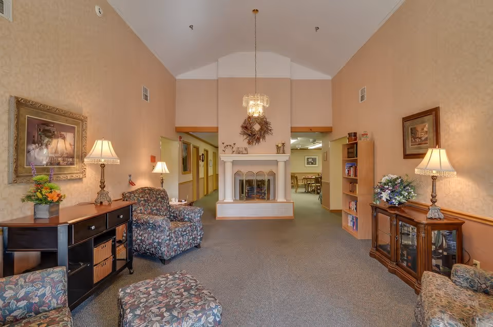 A cozy senior living facility common area with floral upholstered armchairs and ottoman, two wooden side tables with lamps and flower arrangements, a white fireplace with a decorative wreath above it, a bookshelf, and framed artwork on the walls. The room has beige wallpaper, carpeted floor, and a chandelier hanging from the vaulted ceiling. A hallway and dining area are visible in the background.