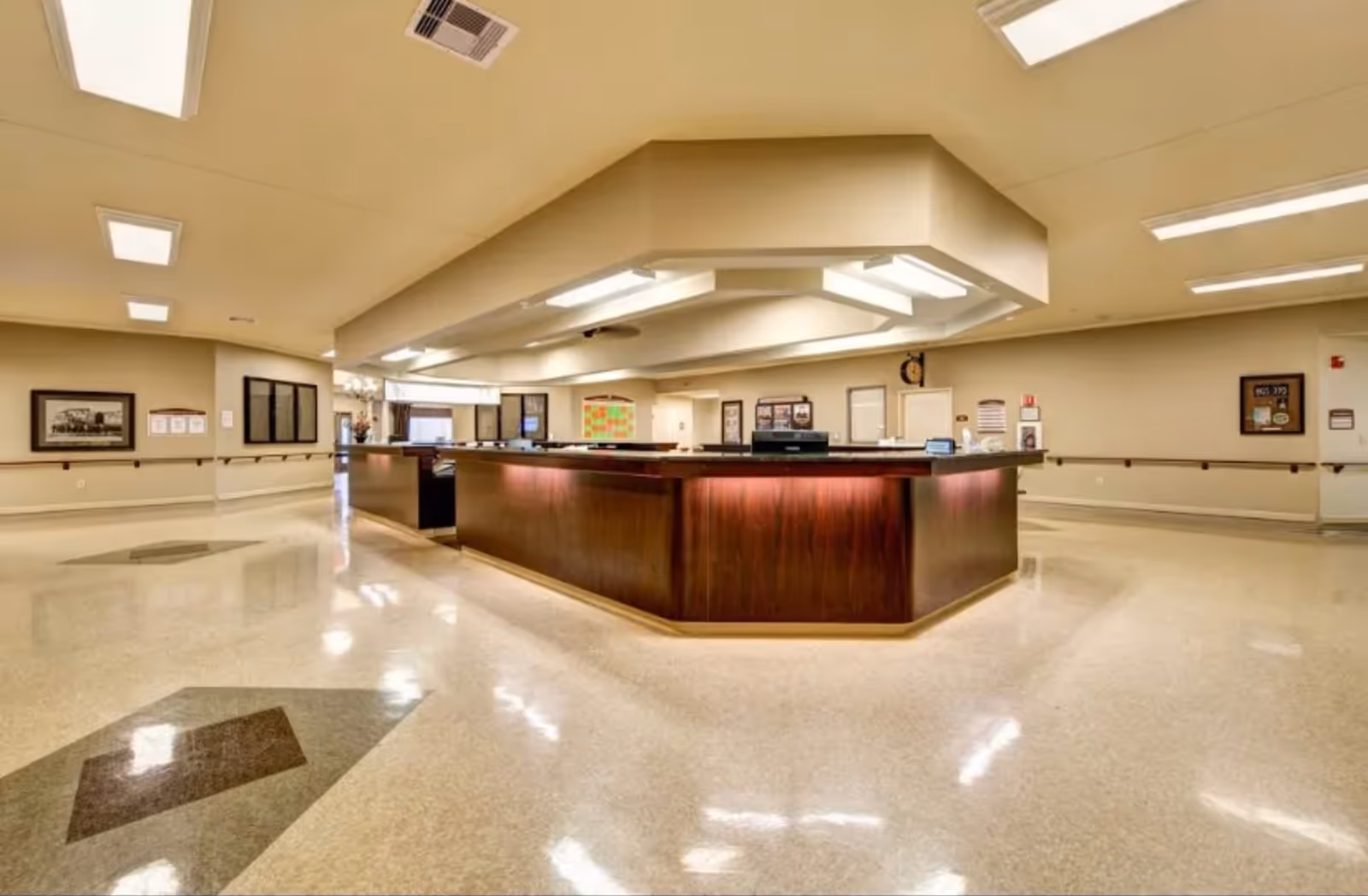 A spacious, well-lit reception or nurse station area inside a healthcare facility with a large wooden counter in the center, beige walls, and polished floors with geometric patterns. The ceiling has multiple fluorescent light fixtures, and there are framed pictures and notices on the walls.