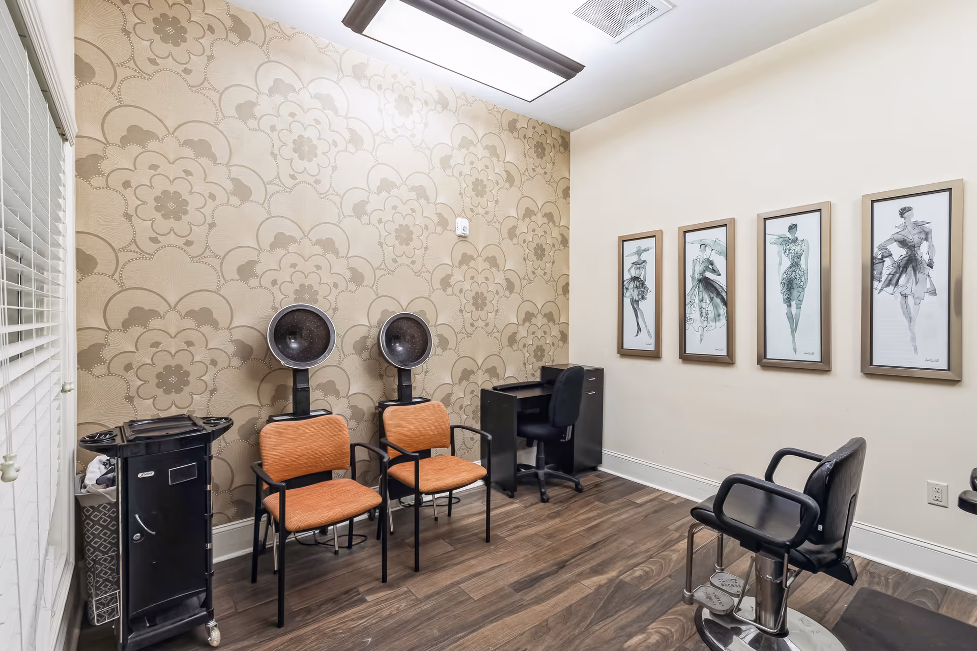 Interior view of a salon room with two orange chairs under hair dryers, a black salon chair, a small black desk with a black office chair, and four framed fashion sketches on the wall. The room has floral patterned wallpaper on one wall and wood flooring.