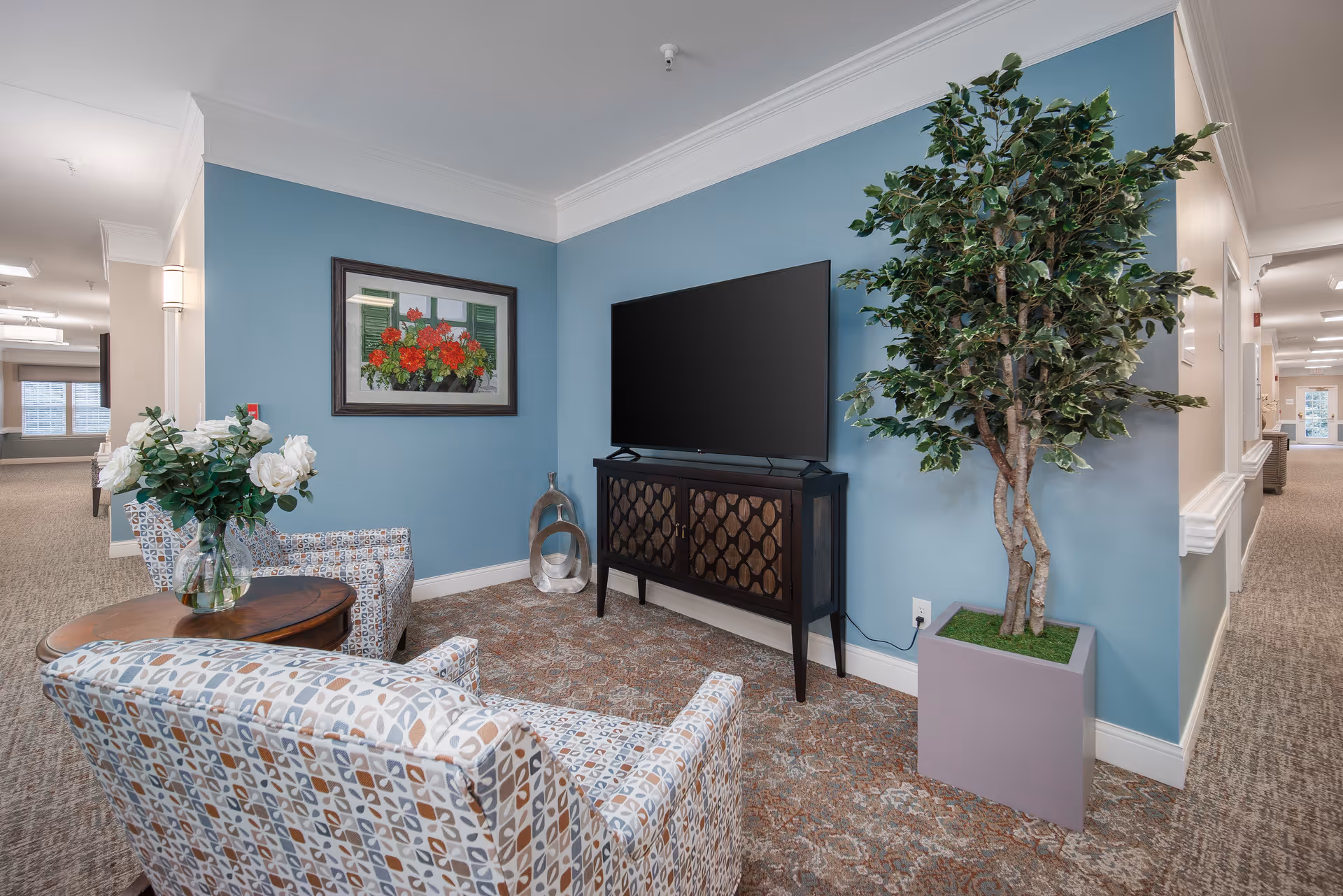 A cozy seating area in a senior living facility with two patterned armchairs, a small round wooden table with a vase of white flowers, a flat-screen TV on a dark wooden cabinet, a large potted artificial tree, and a framed painting of red flowers on a blue wall. The area is carpeted and located in a hallway with soft lighting.