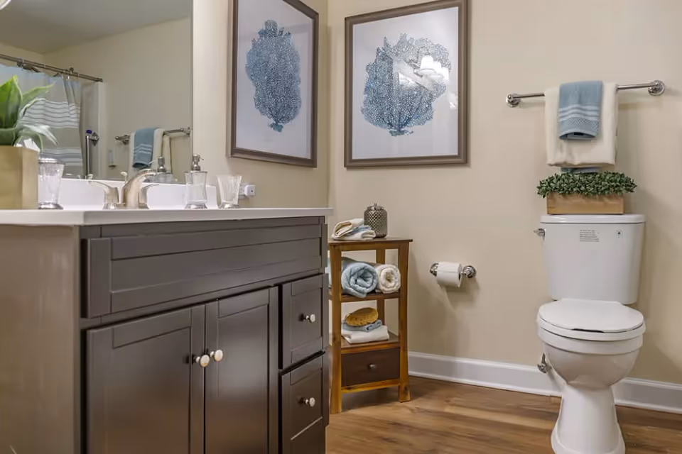 A clean and modern bathroom featuring a dark wood vanity with a white countertop and a sink. Above the vanity is a large mirror. On the right side, there is a white toilet with a small green plant on top and a towel rack above it holding two folded towels. Next to the toilet is a small wooden shelf with rolled towels and bathroom accessories. The walls are light-colored and decorated with two framed blue coral prints. The floor is wood-style.