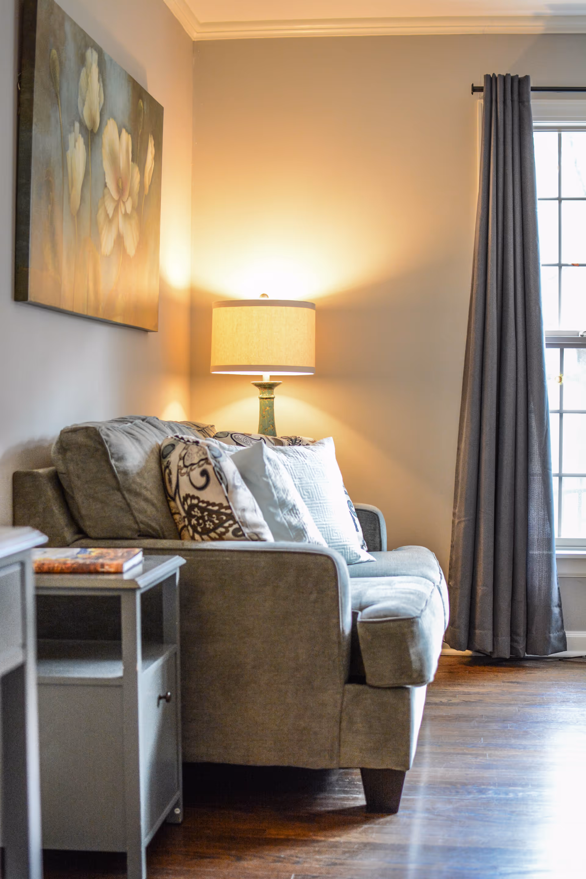 A cozy living room corner with a gray upholstered sofa adorned with patterned and solid pillows. A lit table lamp with a beige shade sits behind the sofa, casting a warm glow on the light-colored wall. A floral painting hangs above the sofa, and a window with gray curtains allows natural light to enter the room. A small gray side table with a book on top is positioned next to the sofa on a wooden floor.