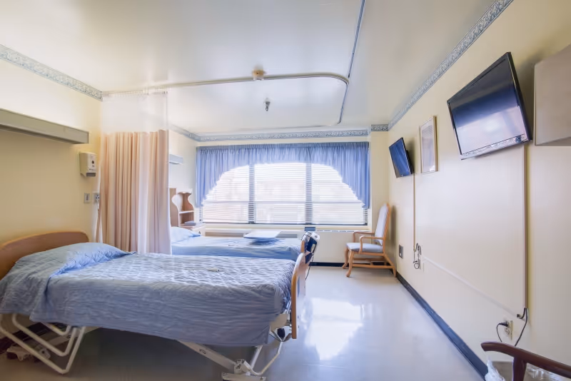 A bright nursing home bedroom with two hospital-style beds covered in blue bedspreads, a privacy curtain between the beds, a large window with blue curtains, two wall-mounted televisions, and a wooden chair near the window.