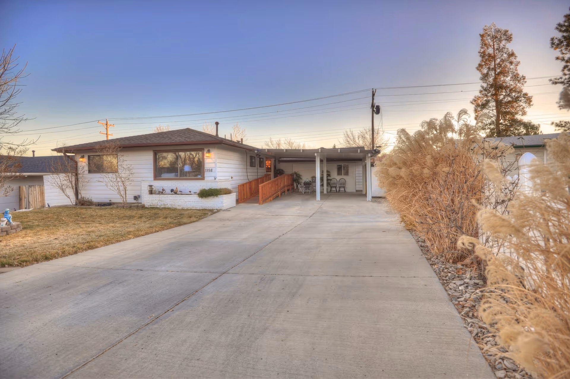 Single-story white house with a long concrete driveway, carport, and ornamental grasses along the right side.