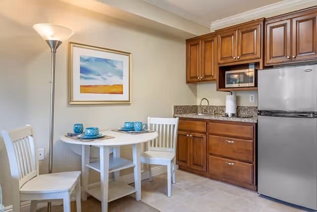 Small kitchen area with wooden cabinets, a stainless steel refrigerator, microwave, and granite countertop with a sink. Next to the kitchen is a small round white dining table set with blue cups and plates, accompanied by two white chairs. A floor lamp and a framed abstract painting hang on the beige wall behind the table.