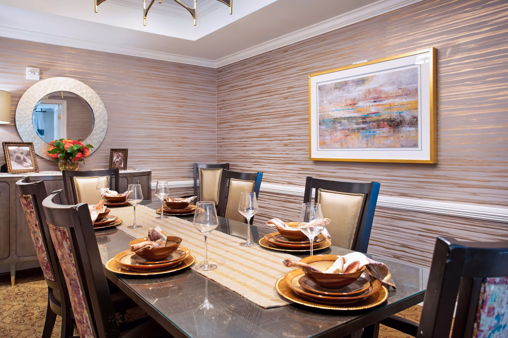 A dining room with a long glass table set for six people. Each place setting includes a brown bowl, a gold charger plate, a folded napkin, and a wine glass. The chairs have dark wooden frames with beige cushioned backs. The walls have a textured beige and brown striped wallpaper. A sideboard with a round mirror, framed photos, and a vase of flowers is visible in the background. A colorful abstract painting hangs on the wall.