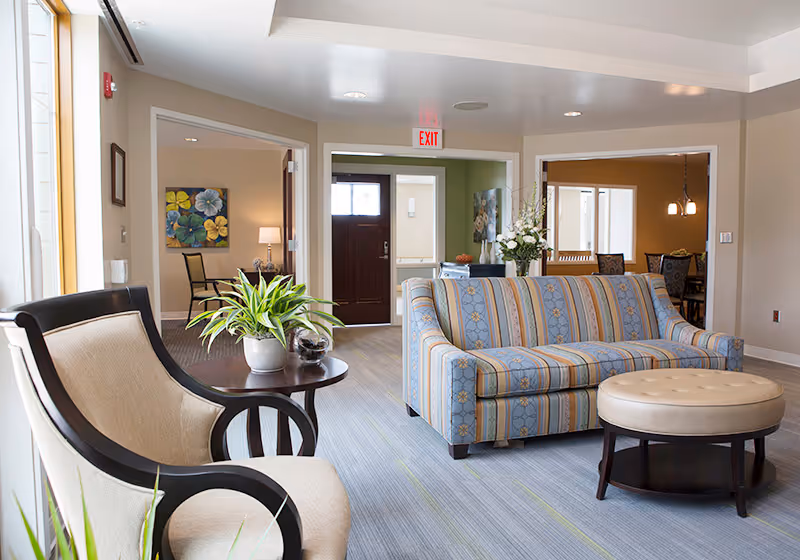 A bright and welcoming living room area in a senior living facility with a striped blue and beige sofa, a round beige ottoman, and a beige armchair with dark wooden arms. A small round table with a potted plant is placed between the armchair and sofa. The room has light-colored walls and carpeted flooring. In the background, there is a dark wooden front door, an exit sign above it, and adjoining rooms with dining tables and chairs visible.