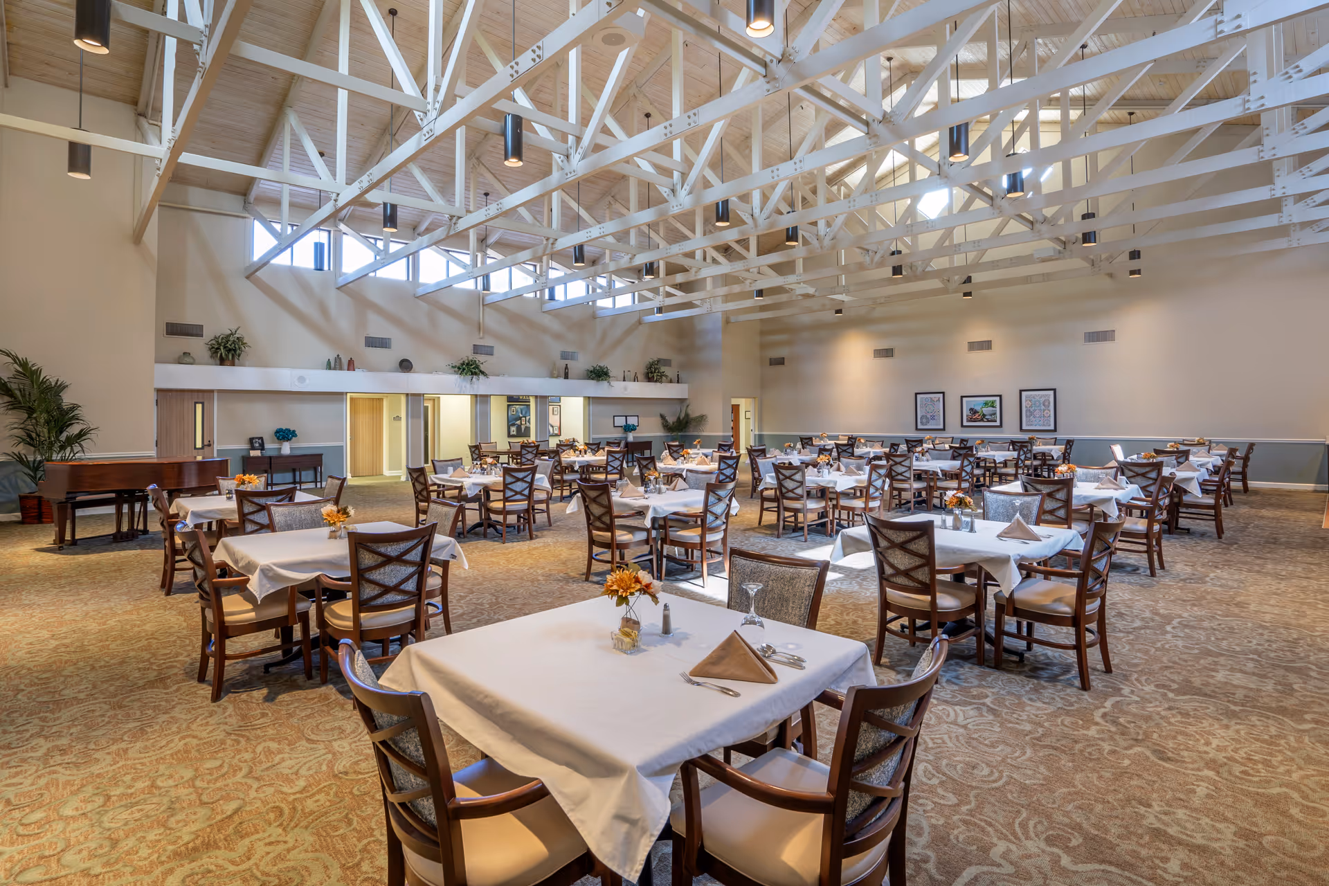 Spacious dining room with multiple tables covered in white tablecloths, each set with napkins, silverware, and small floral centerpieces. The room features high ceilings with exposed white beams and hanging lights, carpeted floor with a patterned design, and a grand piano in the corner. Walls are decorated with framed artwork and plants.