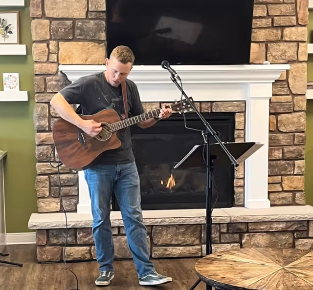 A man plays an acoustic guitar and sings into a microphone in front of a stone fireplace and TV in a communal living room.