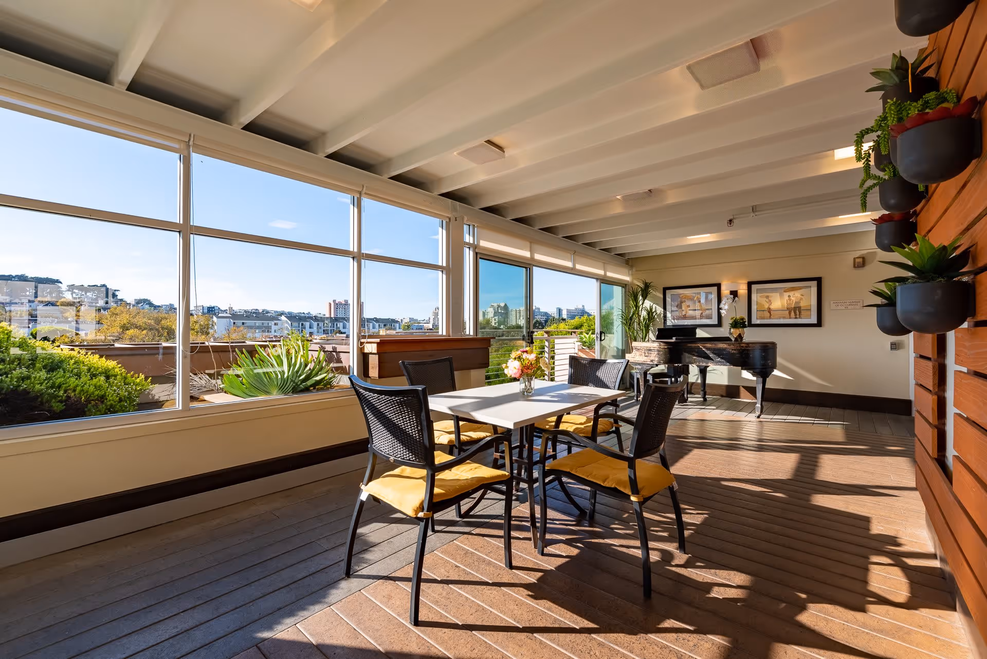 Sunlit indoor lounge with a table and chairs, a grand piano, large windows overlooking the city, and potted plants.