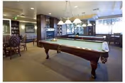 A well-lit interior common room with a pool table in the center, seating areas, and large windows.