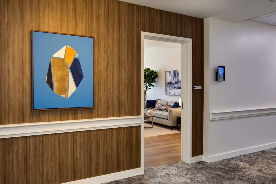 View of a hallway with wood-paneled walls and a framed abstract painting. An open doorway reveals a living room with a beige sofa, decorative pillows, a large plant, and a wall painting.