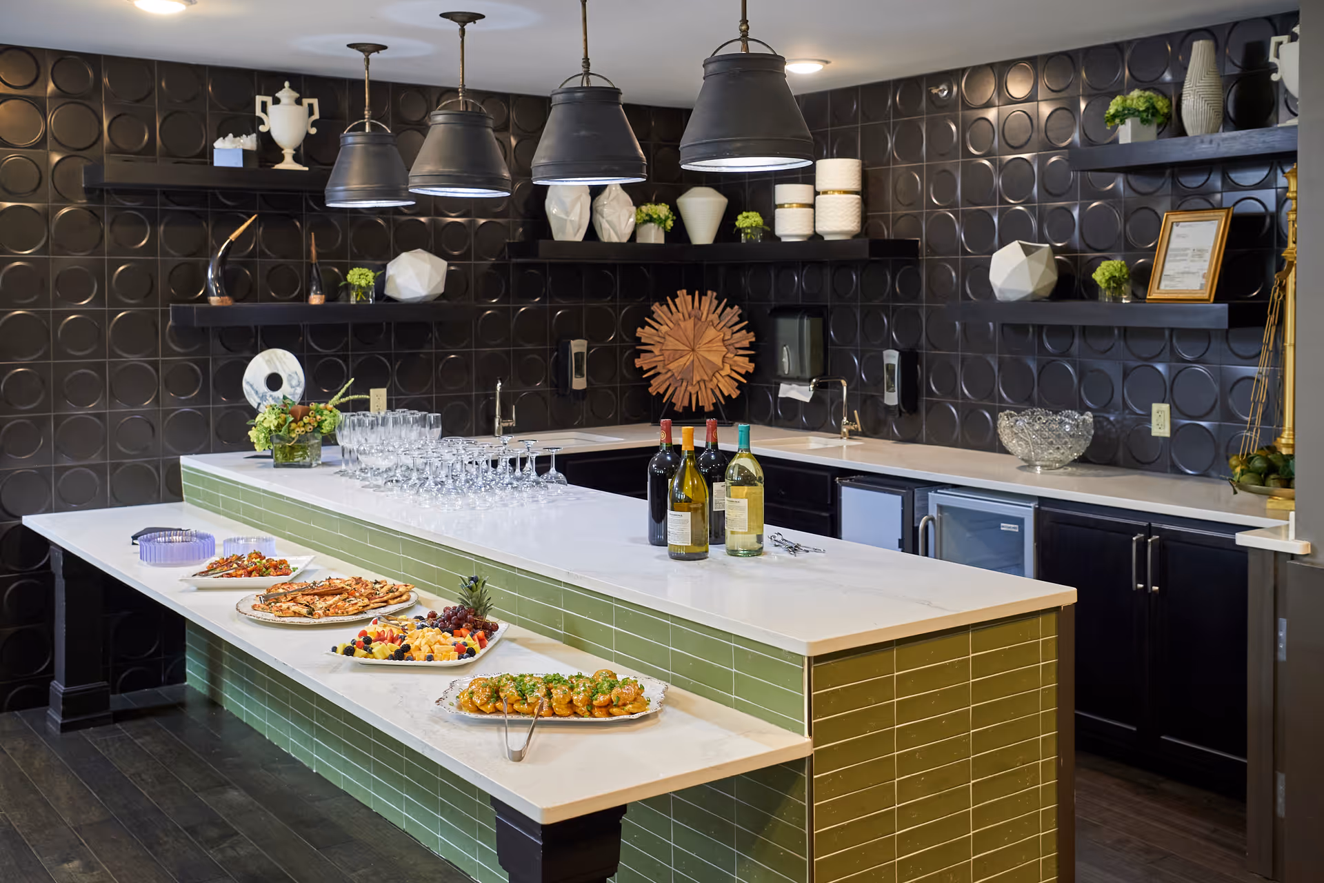 A modern kitchen area with a large island featuring a green tiled base and white countertop. The island is set with plates of appetizers, a fruit and cheese platter, wine bottles, and multiple wine glasses. The background shows black cabinetry, a black textured wall with circular patterns, floating shelves with decorative items, and three large pendant lights hanging above the island.