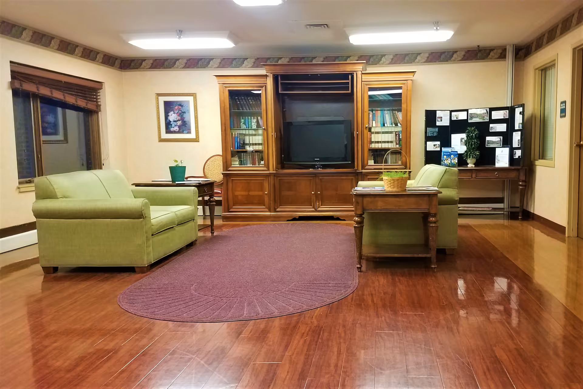 A cozy common area with two green armchairs facing a wooden entertainment center with a television and bookshelves. There is a purple rug on the wooden floor, a small wooden table with a basket and magazines, and a side table with a potted plant. The walls are decorated with framed pictures and a bulletin board with various notices.