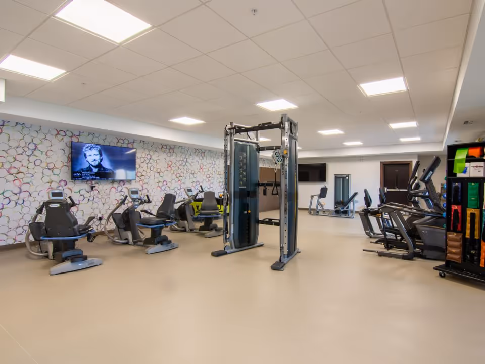 Bright fitness room with multiple cardio machines, a central weight station, and a wall-mounted TV.